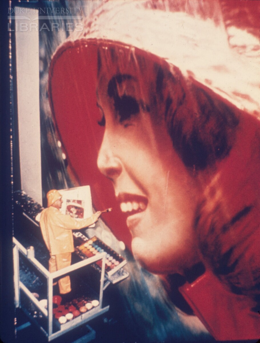 Artist in rain gear with a paint brush in front of billboard 1960s